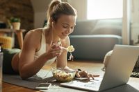 young-happy-sportswoman-eating-salad-using-laptop-while-relaxing-floor-home